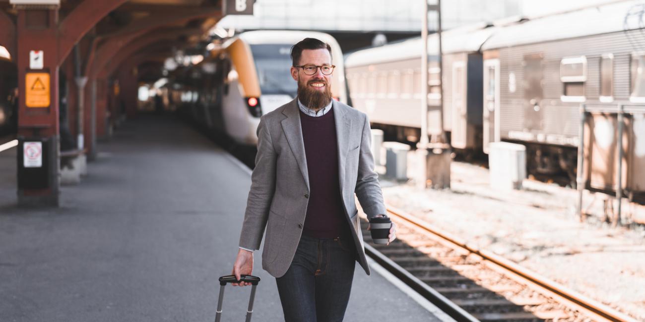 A smiling man with a suitcase walks along a train station platform in Scandinavia.