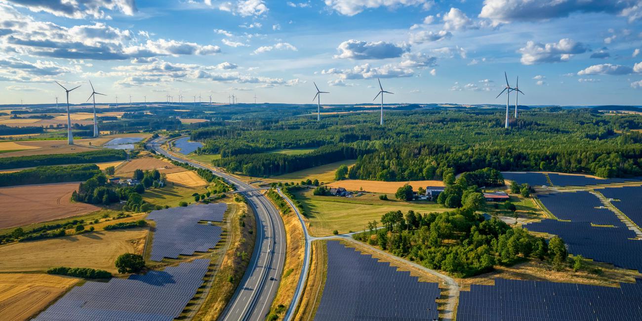 Aerial view of solar power station on either side of a highway with wind turbines sticking out above a forest in the background.