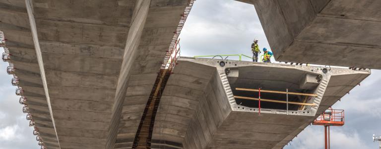 Two construction workers stand on a concrete bridge under construction