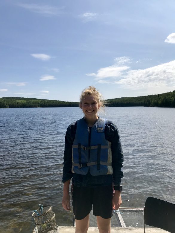 A woman wearing a lifejacket stands smiling on the dock of a lake. 