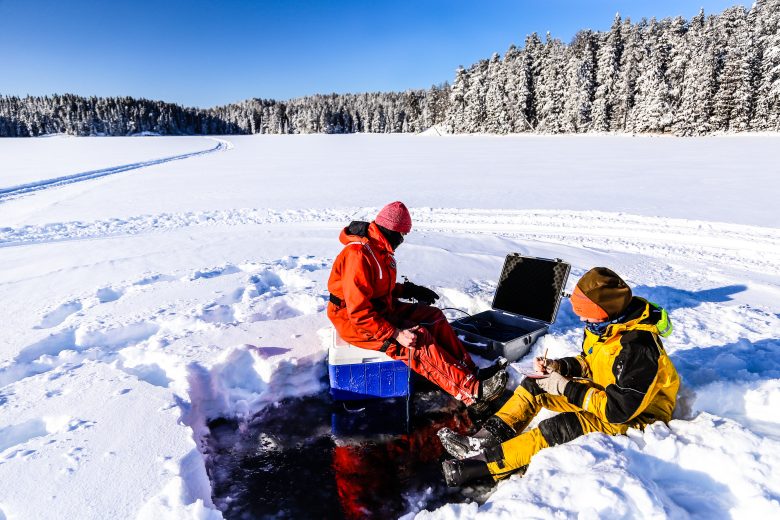 Scientists look at a computer while sitting on a frozen lake covered in snow in Canada's boreal forest.
