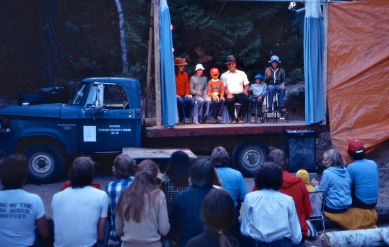 Group of people with back to camera watch people on a stage on a trailer sitting in a straight line