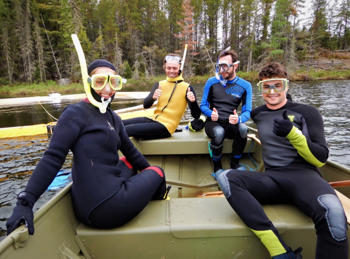 Four people sit on a boat with black and yellow scubadiving equipment