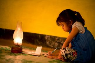 A young girl sits by a lamp on the floor writing on a piece of paper.