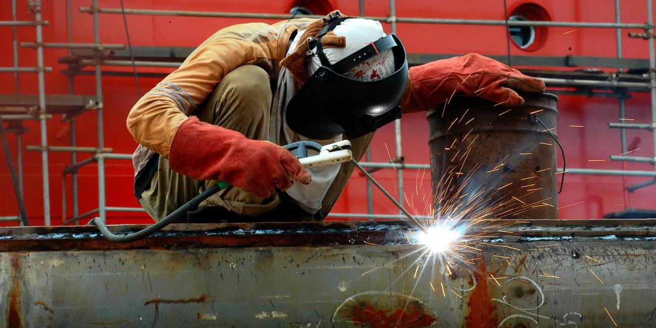 A worker wears a mask while he is welding in a bright red industrial background.