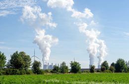 A power plant and wind turbines operate near a green field.