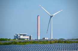 Solar panels, a wind turbine, and coal station are seen against a blue sky.