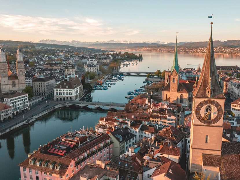 A clocktower oversees town buildings next to a lake and mountains.