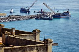 Cranes on ships are used to pile rocks along a road over water.