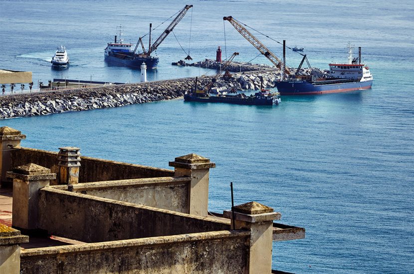 Cranes on ships are used to pile rocks along a road over water.
