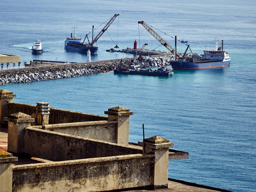 Cranes on ships are used to pile rocks along a road over water.