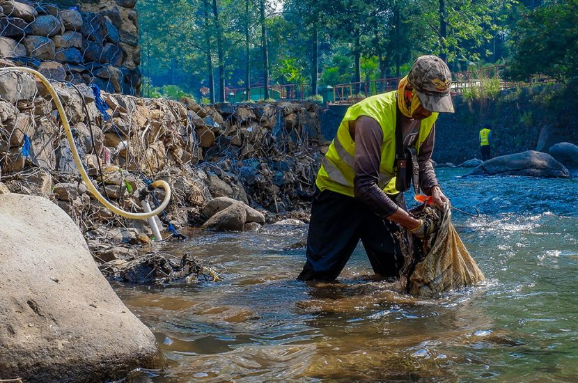 A person in a safety vest places items into a sack, standing in a narrow river.