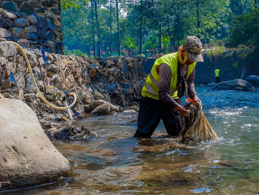 A person in a safety vest places items into a sack, standing in a narrow river.