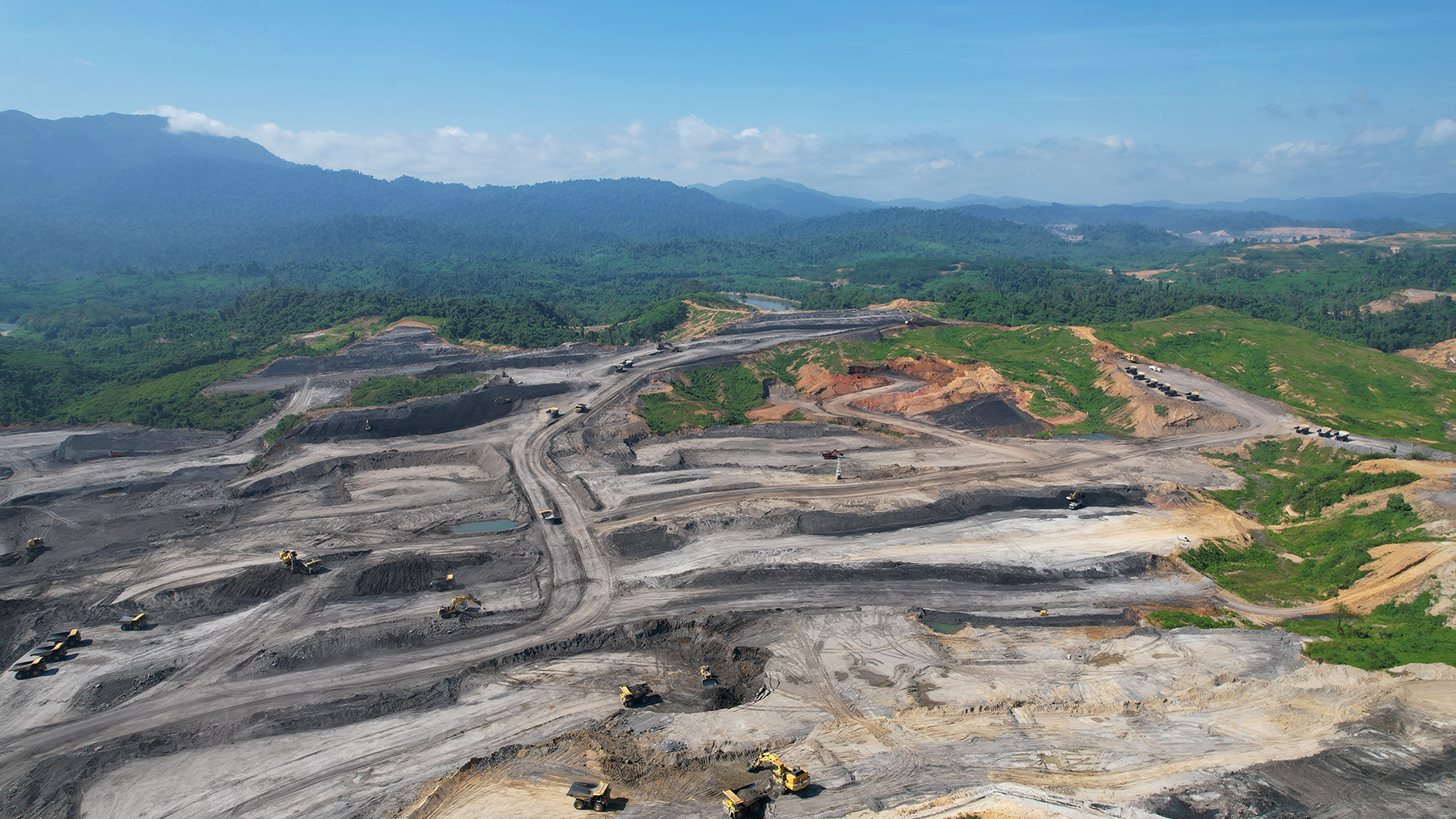 High level view of machinery in an open pit coal mine with trees and green mountains in the background.