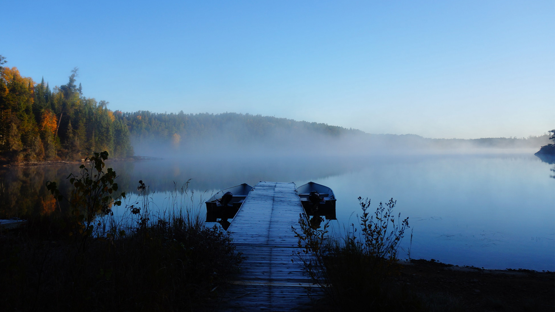 Two empty boats sit on either side of a dock as it extends into a tree-lined lake on a frosty morning.
