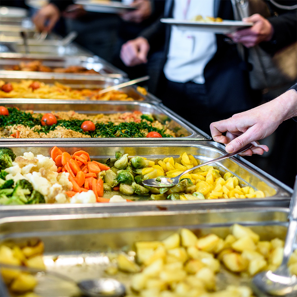 Close up of trays of healthy food in a buffet line with people out of focus in the background.