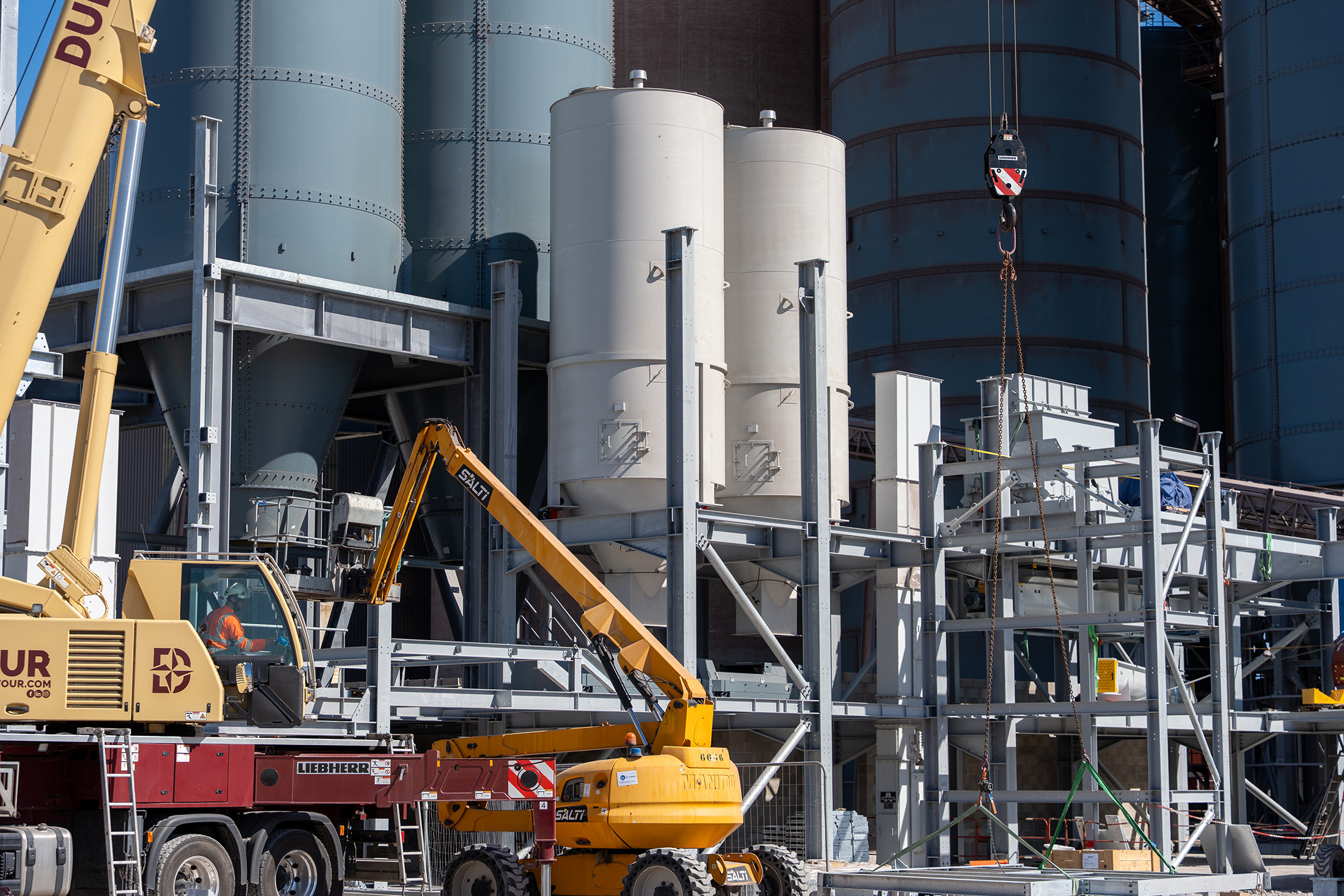 Yellow construction trucks in front of grey steel beams and tanks.