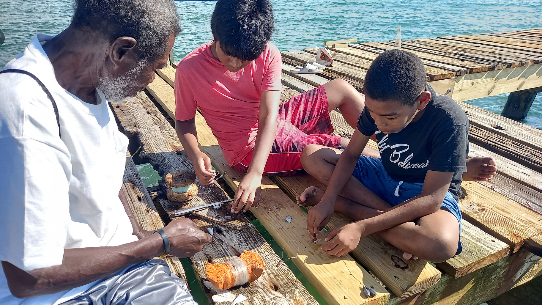 A grey-haired man sits with two children on a dock, showing them his fishing tools.