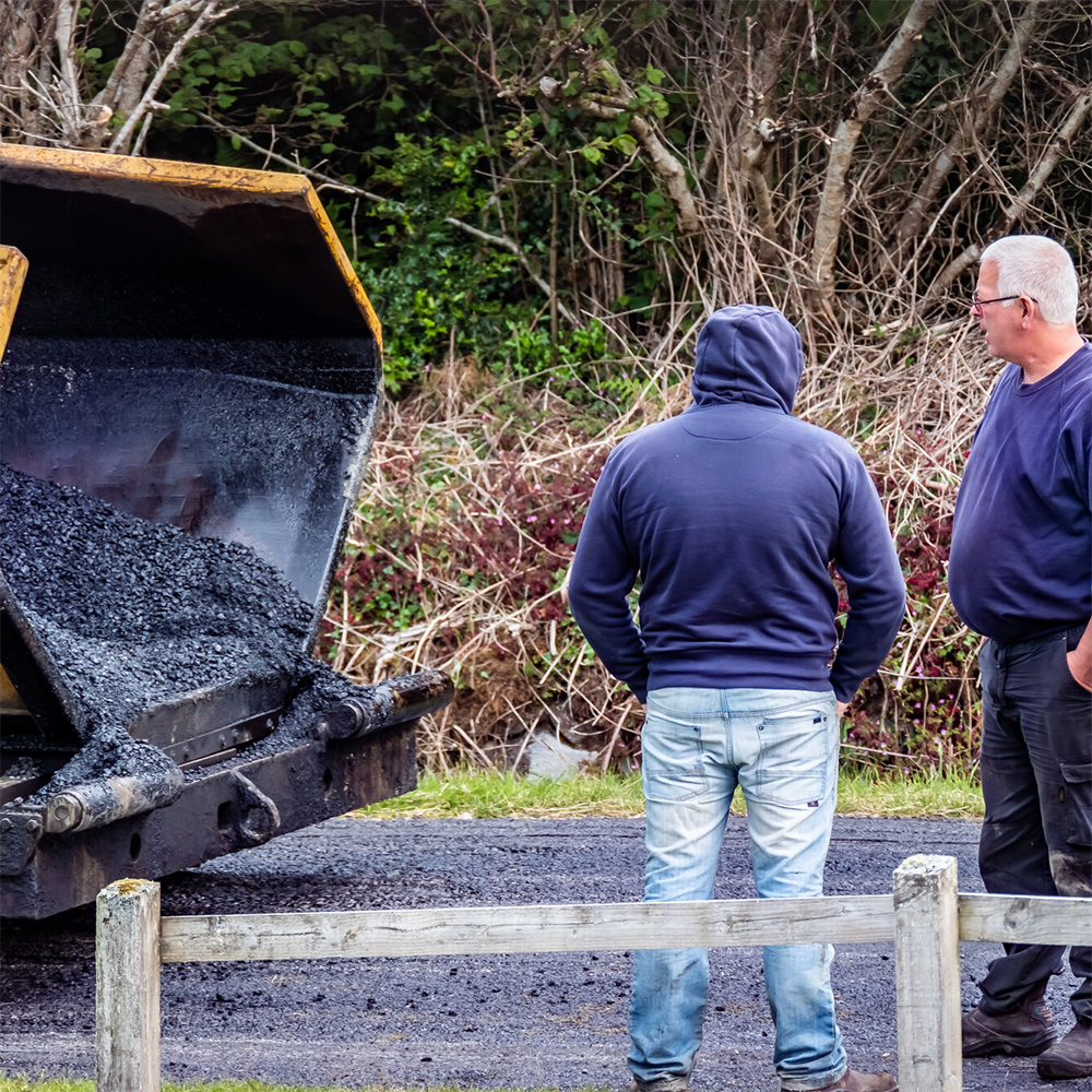 Two construction workers stand behind a truck with asphalt as they pave the road.