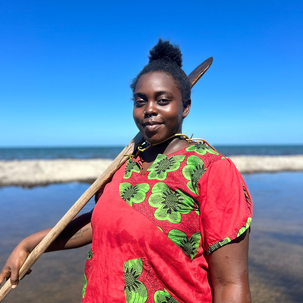 Black woman in focus with blurry coastal ocean behind her. She is wearing a red dress with green flowers.