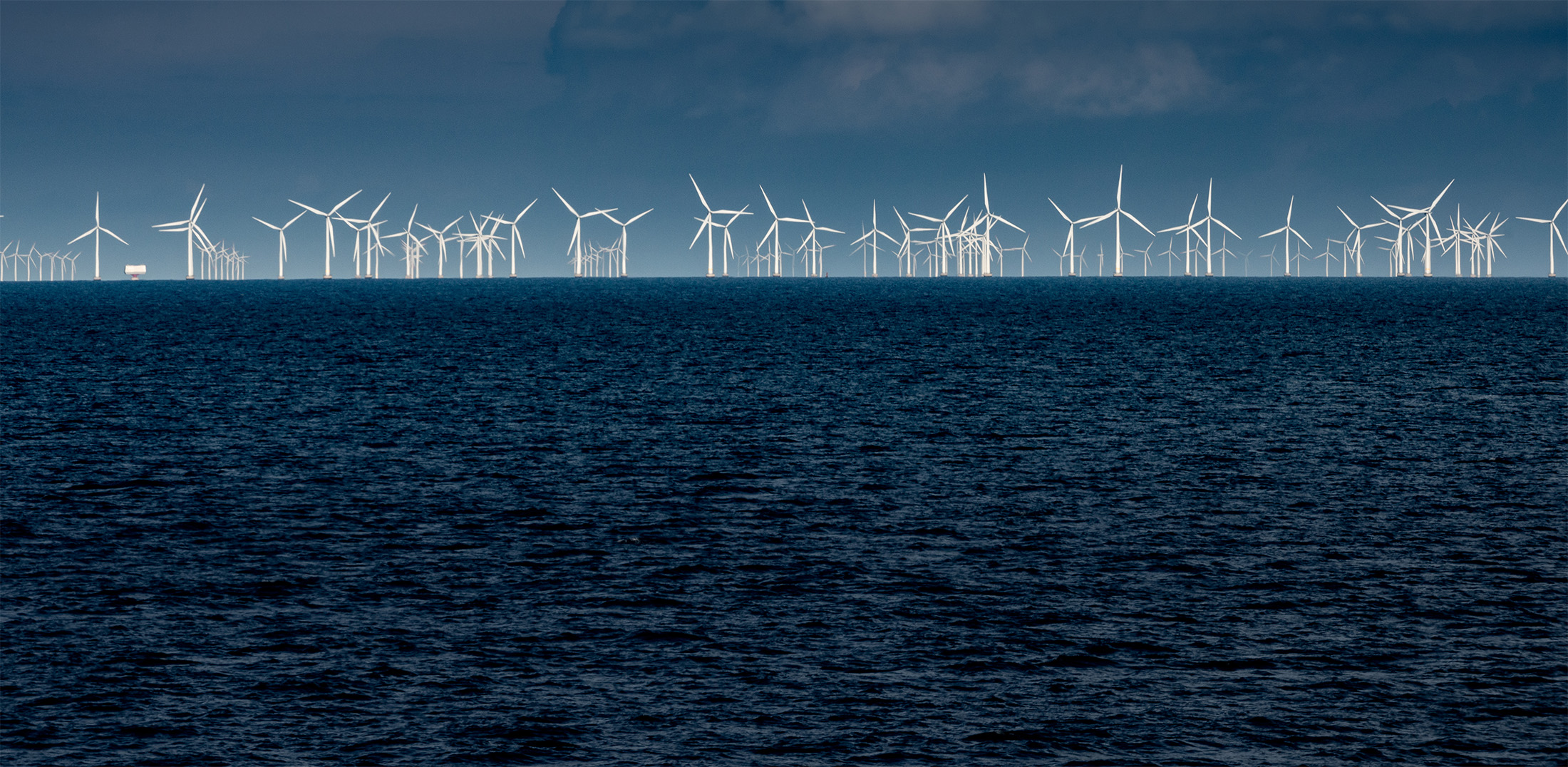 Line of wind turbines along the horizon with ocean in foreground.
