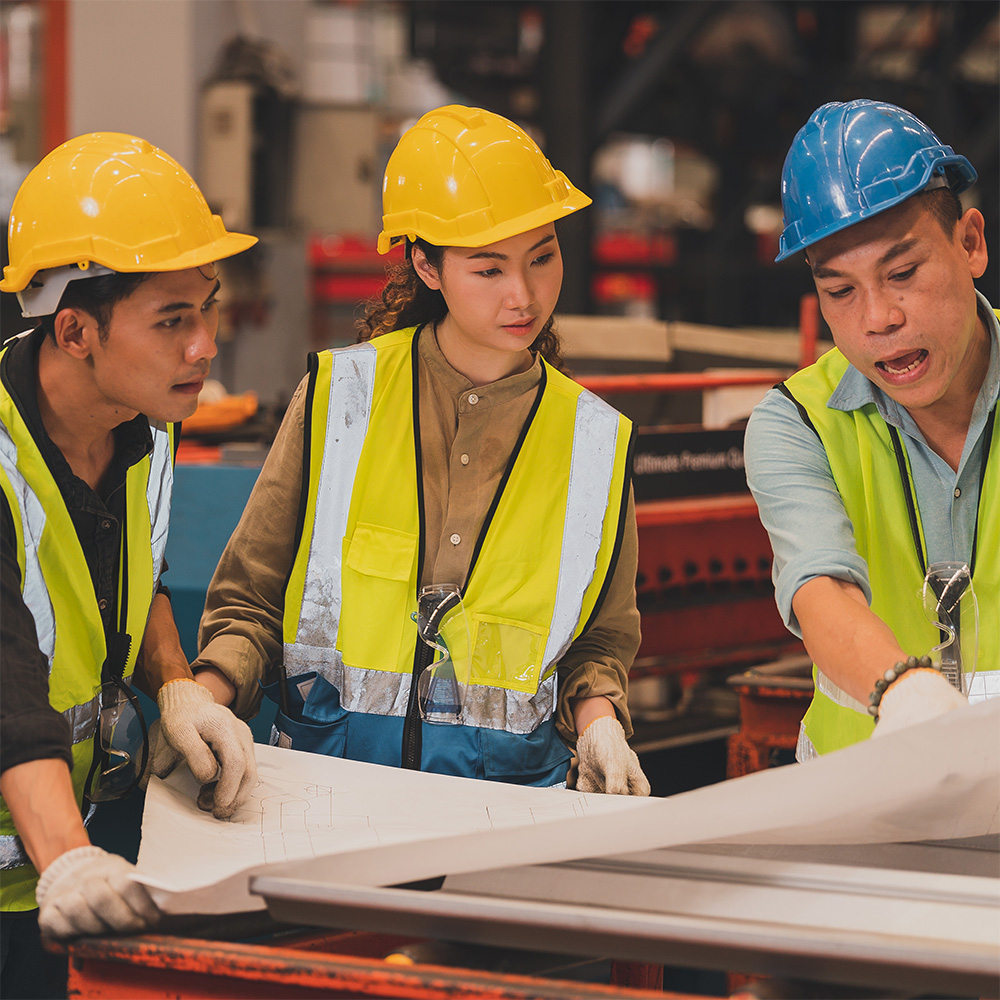 Three people wearing heard hats and hi vis vests review construction plans on a large piece of paper on a table.