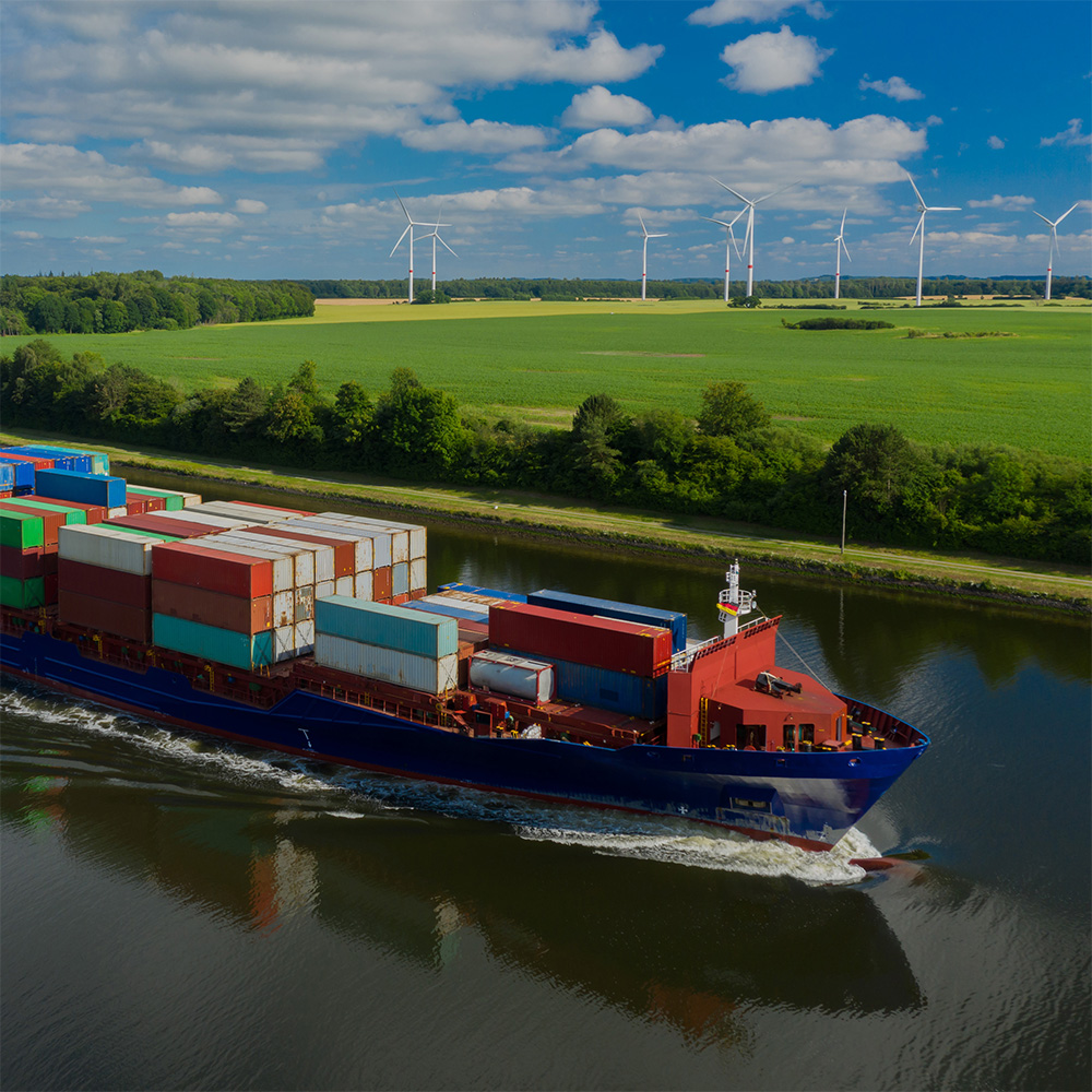 Freight ship travels down river in foreground with green fields and wind turbines in the background.