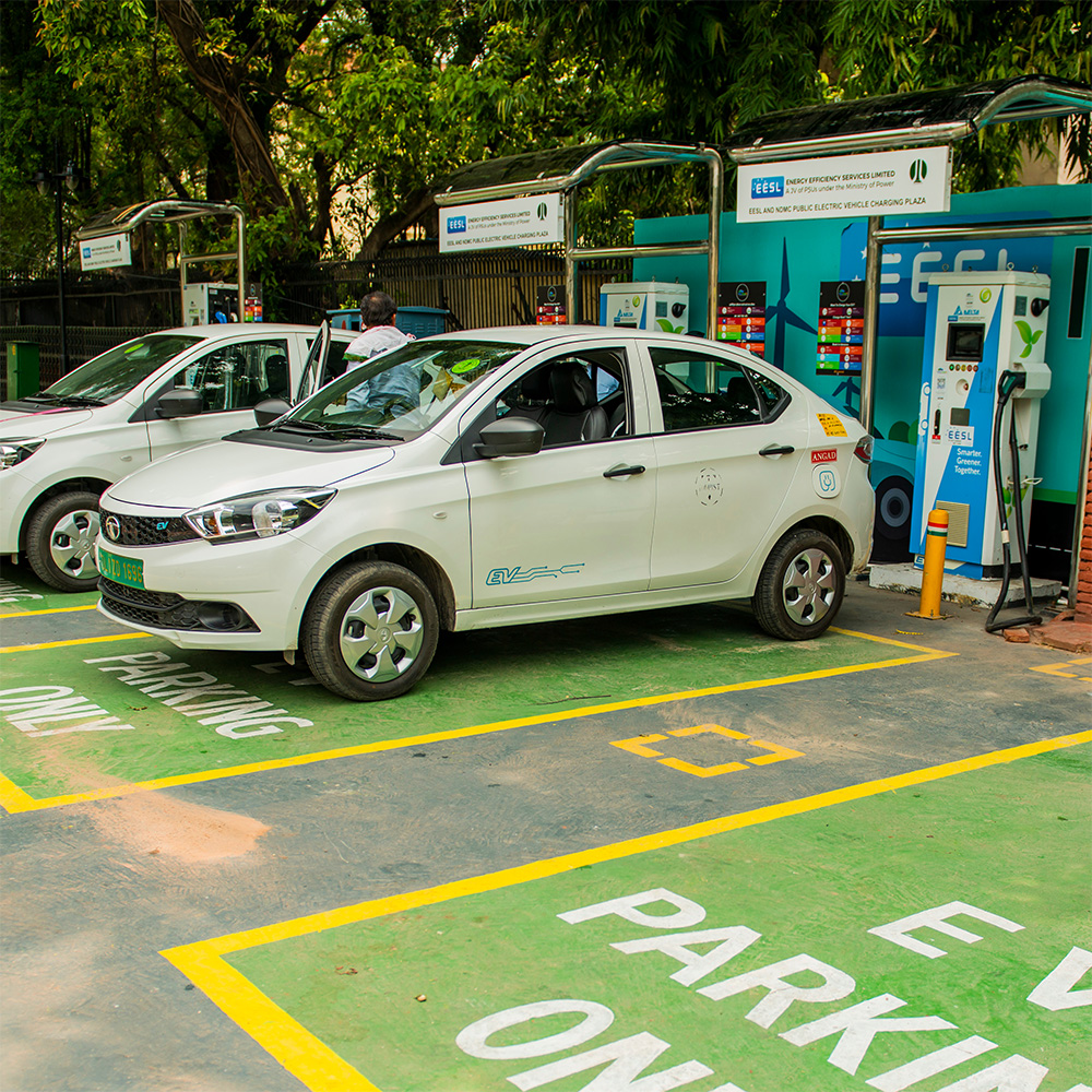 Two white cars sit at an EV charging station with trees behind.