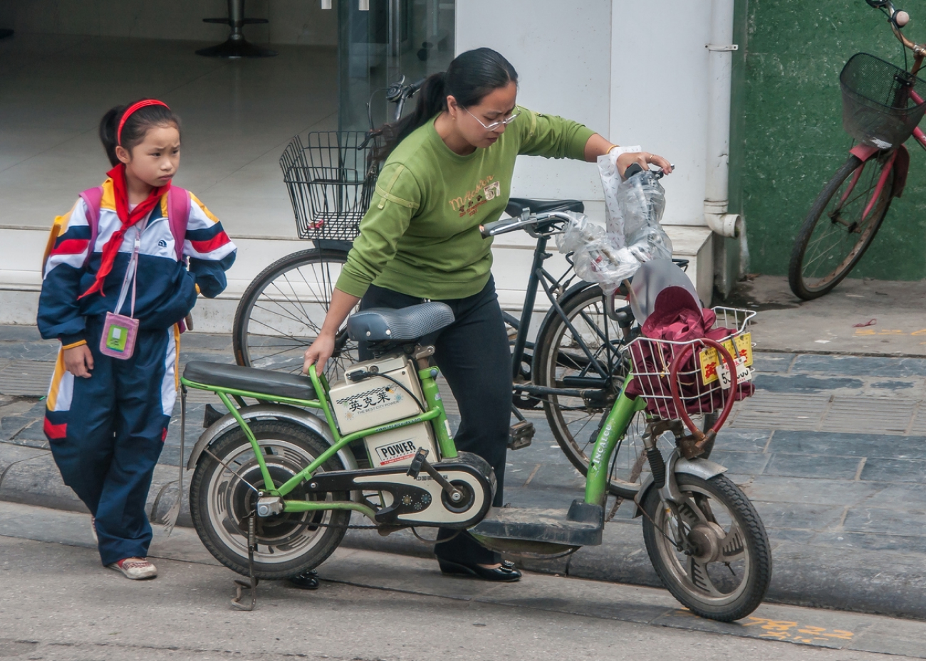 Mother in green-black garb and kid-daughter in multicolor clothing with green-gray electric bike on street.