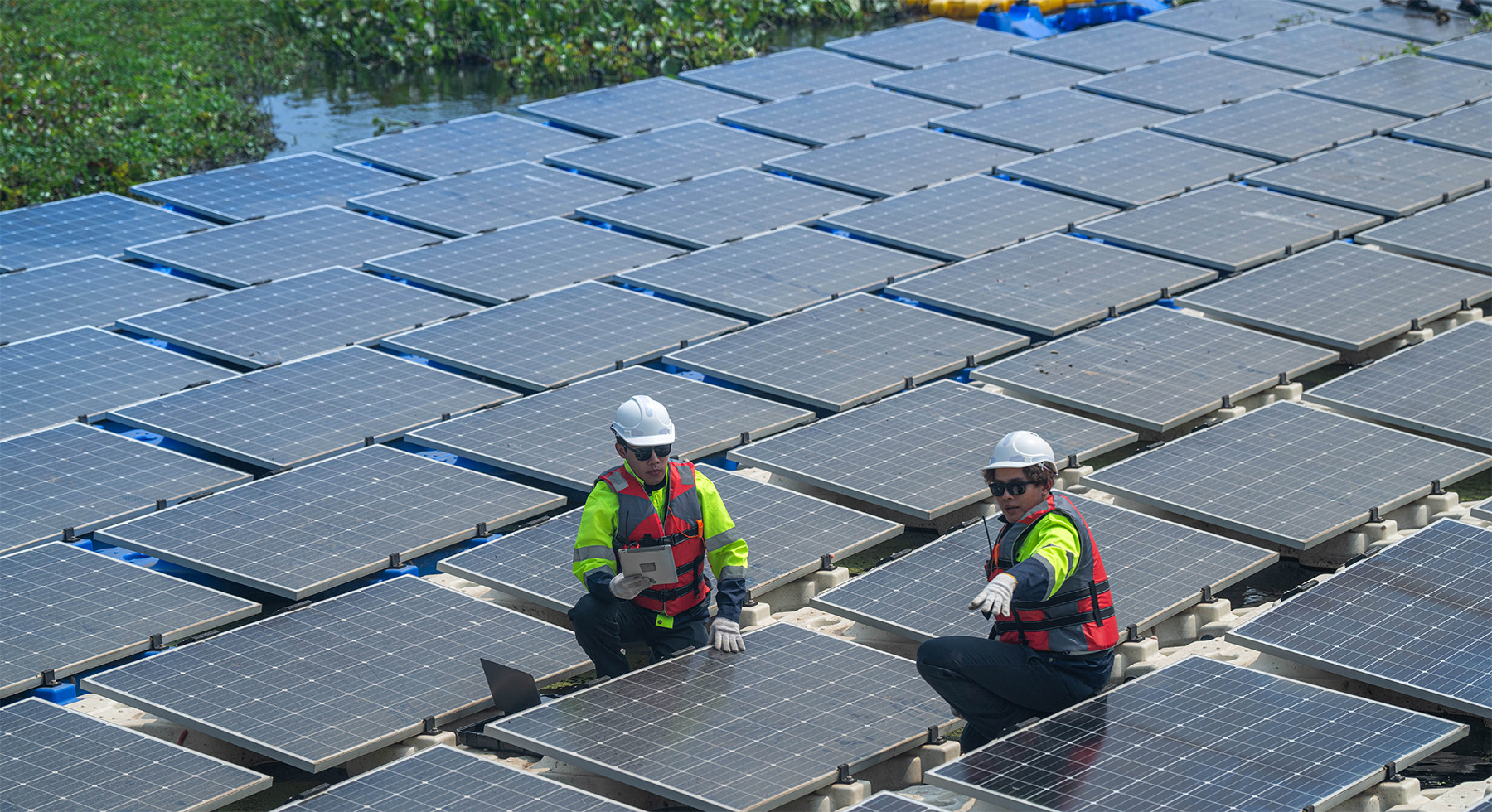 Two engineers crouch next to solar panels next to river.