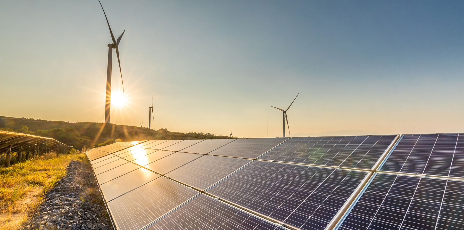 Solar panel in foreground, wind turbine on hill in midground with setting sun behind it.