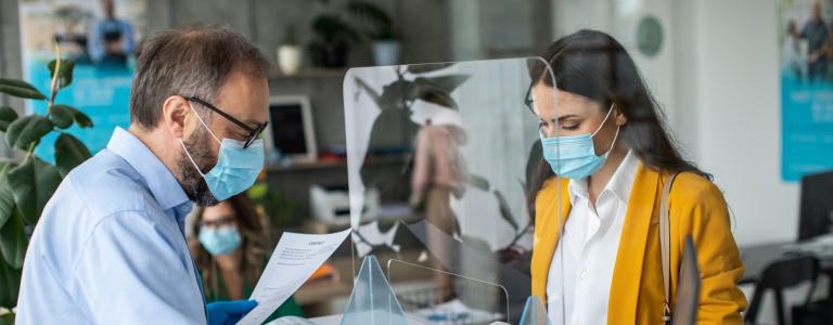 A man and woman wearing masks and gloves look at paperwork with plexiglass in between them