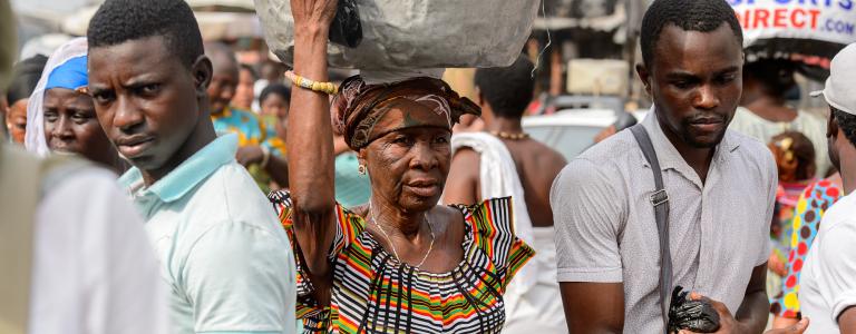 A woman carries a large sack on her head while walking through a busy market, flanked by two men