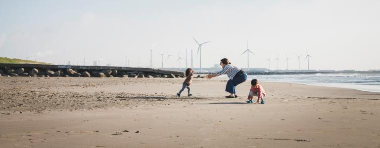 A mother and her two small children play on a beach with wind turbines in the distance