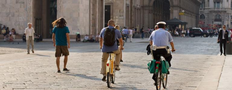 Two cyclists ride along a quiet street in Milan, Italy, seen from behind