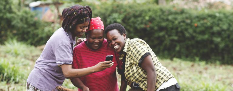 Three women on a farm in Kenya look at a mobile phone while smiling as a small child stands beneath