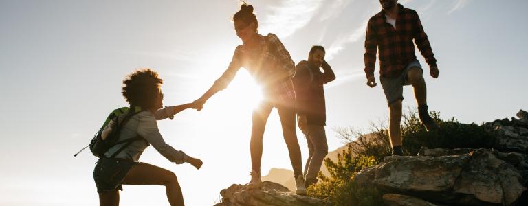 four people climb a hill at sunset, with one helping to pull another up