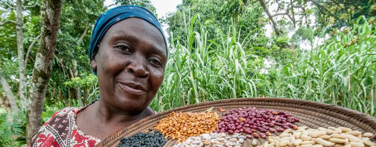 An African woman holds up a tray of various beans and looks at the camera