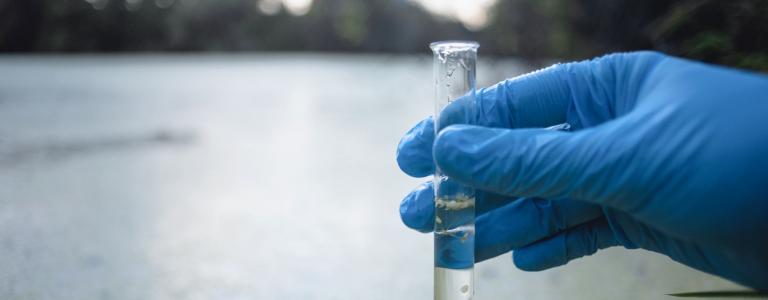 A hand in a blue latex glove holding a test tube in front of a lake