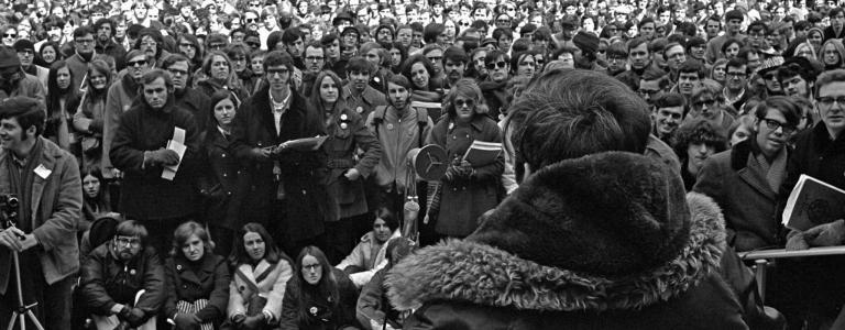 Black and white photo of a large crowd watching as a man speaks from a podium