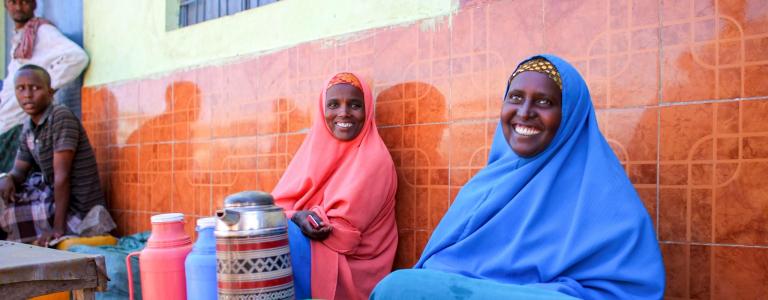 Two Somali women in colourful traditional dress smile at the camera in front of carafes of tea