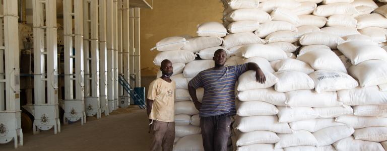 Two men stand next to a large stack of bagged rice in a warehouse