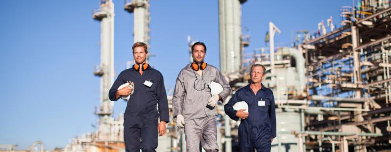 Three oil workers walk away from refinery on sunny day, holding hard hats.