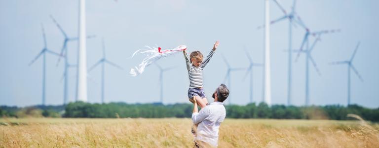 A father holds up his young daughter in a field of wheat with wind turbines in the background