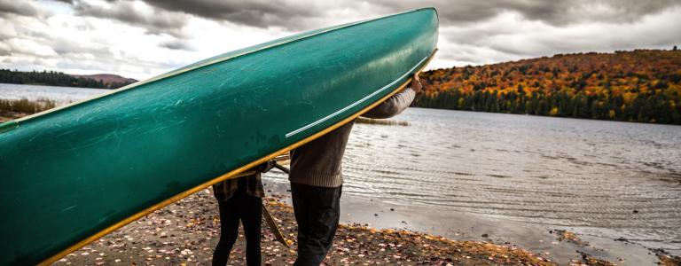 Two people carry a green canoe over their heads toward a lake on a cloudy day
