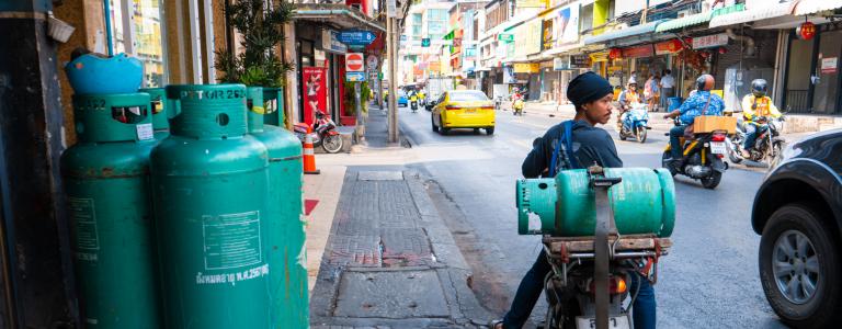 A man sits on a motorbike with a propane gas cylinder strapped to the back on a city street