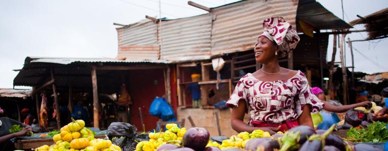 A woman stands behind a vegetable stall outdoors in traditional African dress