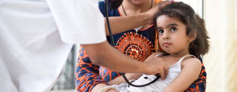 A woman in a health facility in India holds her daughter in her arms as a doctor undertakes a check-up.