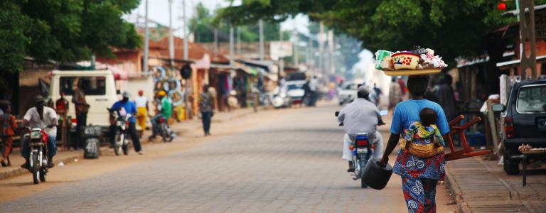Street scene in Benin. A mother carrying a kid and some man riding motorcycles.