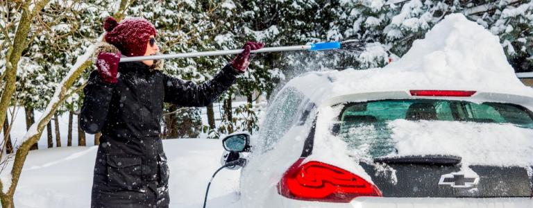 After a snowfall in St-Hugues, Quebec, Canada. An electric car is covered with snow. The car is the Chevrolet Bolt. The vehicle is charging. A lot of snow covers the car. A woman is busy getting the car out of the snow.